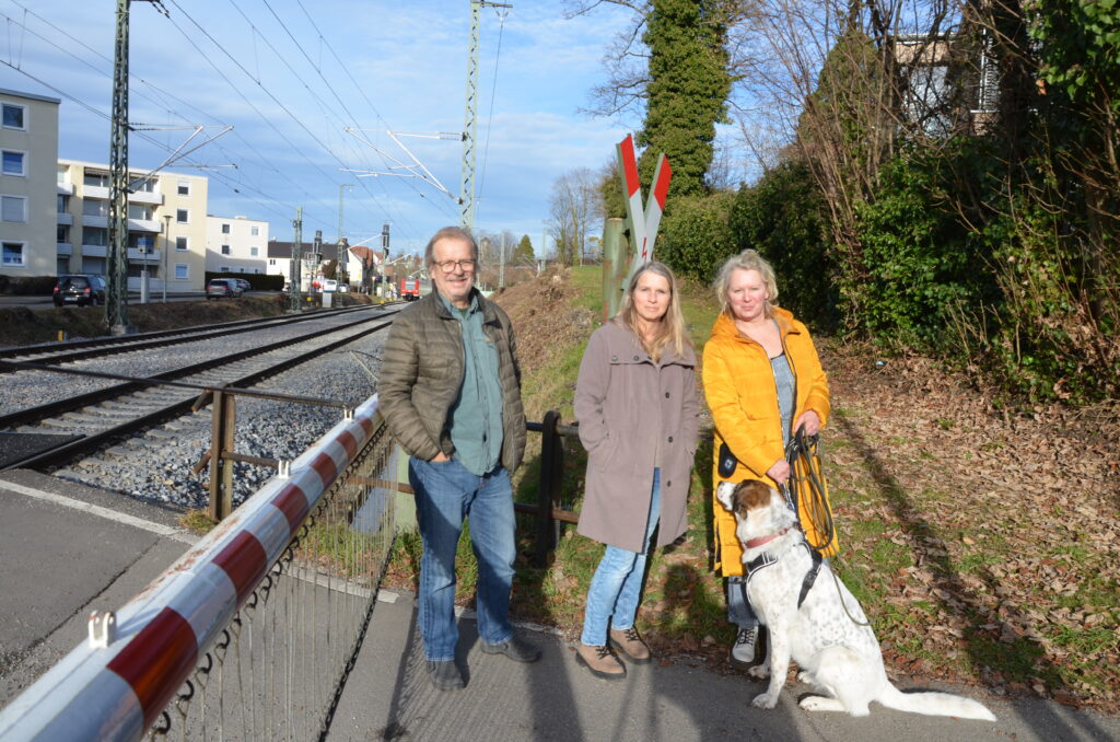 Drei Menschen stehen an einer verschlossenen Bahnschranke.