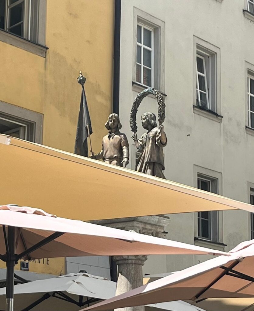 Ein Mädchen und ein Junge die in Festkleidung, als Statuen auf einem Brunnen stehen