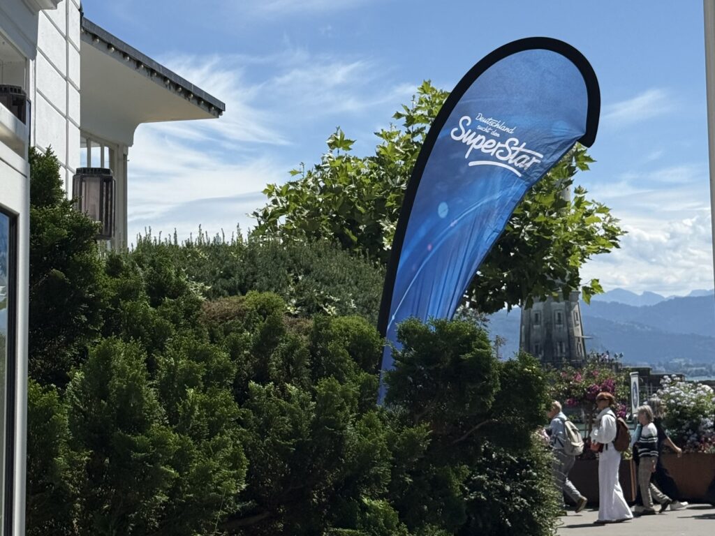 Blick auf ein Banner mit "DSDS"-Schriftzug. Im Hintergrund die Lindauer Promenade und der Bodensee. Links die Hausarbeit eines Hotels