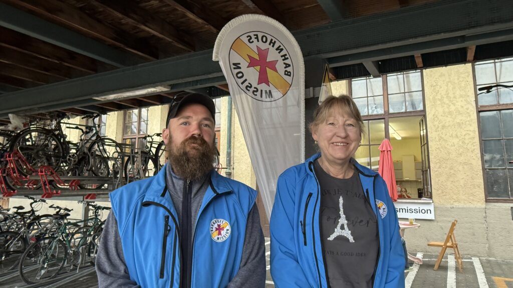 Ein Mann und eine Frau am Bahnhof vor einem Banner mit dem Schriftzug "Bahnhofsmission Lindau". Links von ihnen Fahrradständer, im Hintergrund ein Tisch mit Sitzmöglichkeiten.
