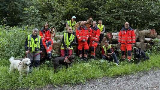 Gruppenfoto im Wald: Im Hintergrund liegen aufeinandergestapelte, gefällte Bäume. Im Vordergrund stehen mehrere Menschen in roter Uniform und mit Warnwesten sowie mehrere Hunde.