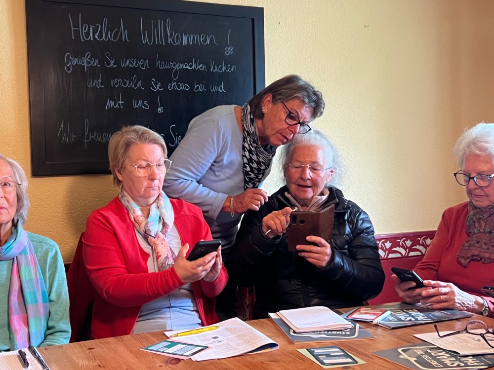 Mehrere seniorinnen sitzen an einem Tisch und haben ein Handy in der Hand.